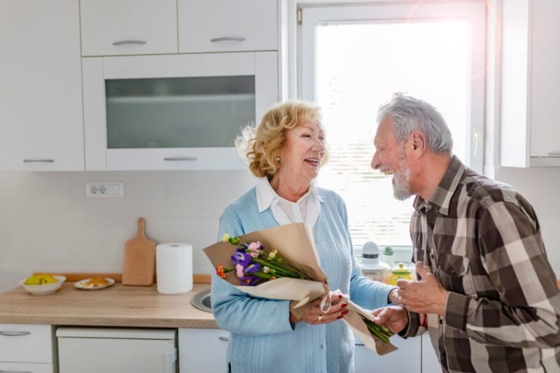 Zeitlose Liebe - Platinhochzeit nach 55 Jahren Ehe. Ein älteres Paar, wo der Mann der Frau lachend einen Blumenstrauß zum Jubiläum überreicht.