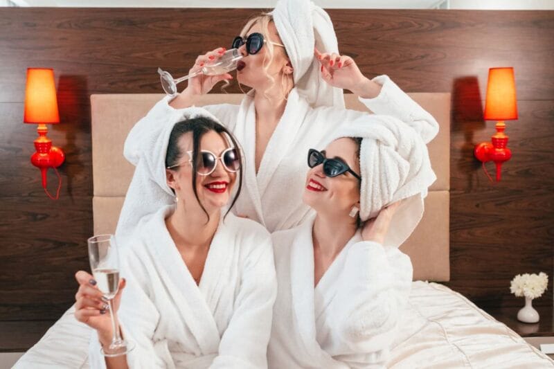 Drei Frauen in weißen Bademänteln mit Champagnerglas bei der Bachelorette Party.