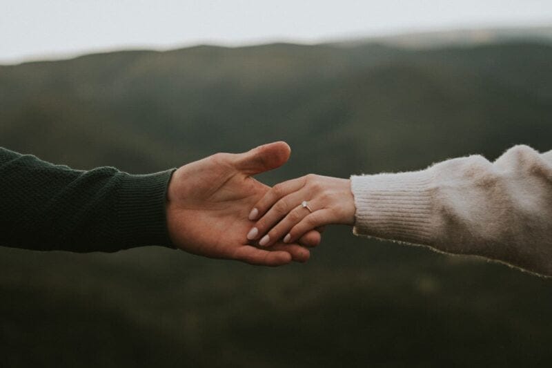 Mann und Frau halten Hände mit Verlobungsring auf der linken Hand der Frau. Landschaft im Hintergrund.