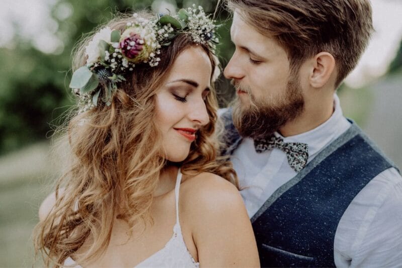 Ein eleganter Mann trägt Jeans und Sakko zur Hochzeit, während eine Braut in einem Boho-Hochzeitskleid neben ihm steht.