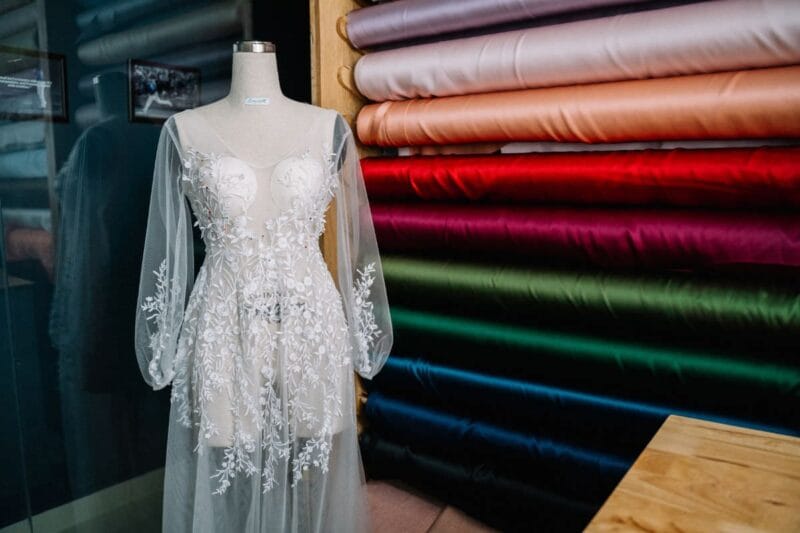 Schaufensterpuppe mit einem bunte Hochzeitskleid vor einer Wand aus Stoffrollen.