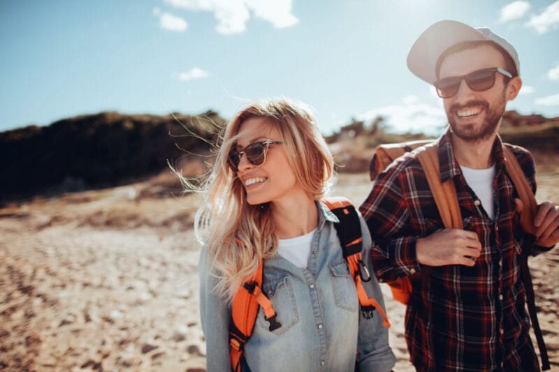 Ein verlobtes Paar genießt gemeinsam einen sonnigen Tag am Strand. Beide tragen Rucksäcke und lachen glücklich. - Wie lange kann man verlobt sein?