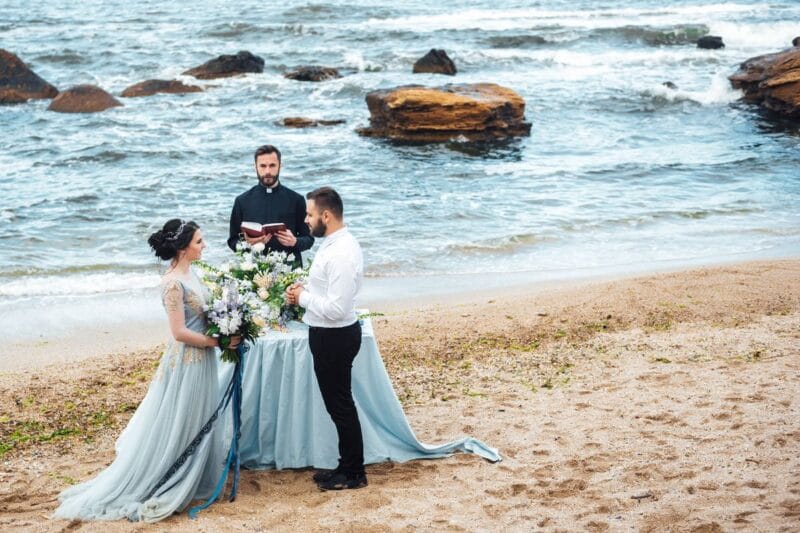 Hochzeitspaar bei einer Zeremonie am Strand, während ein Priester außergewöhnliche Trausprüche aus der Bibel vorliest.