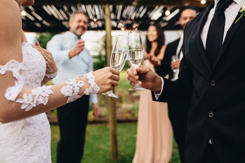 Braut und Bräutigam stoßen mit Sektgläsern auf ihrer Hochzeit an, umgeben von Gästen.