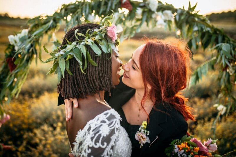 Zwei Frauen geben sich einen Hochzeitskuss vor einem Blumenbogen