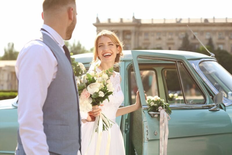 Die lächelnde Braut steht neben einem blauen Hochzeitsauto, wer fährt das Hochzeitsauto?