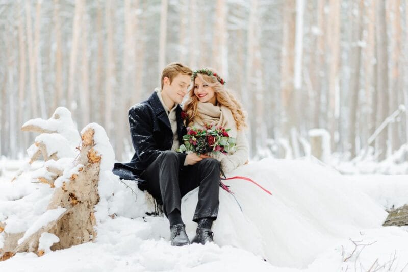 Brautpaar sitzt im Schnee bei einer Winterhochzeit, umgeben von winterlicher Landschaft.
