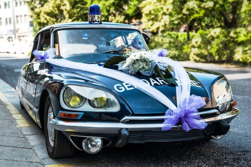 Ein klassisches Polizeiauto, geschmückt mit lila und weißen Autoschleifen für eine Hochzeit, parkend an der Straße.