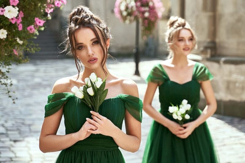 Zwei Brautjungfern in dunkelgrünen Kleidern, eine im Vordergrund hält weiße Blumen, während die andere im Hintergrund verschwommen steht.