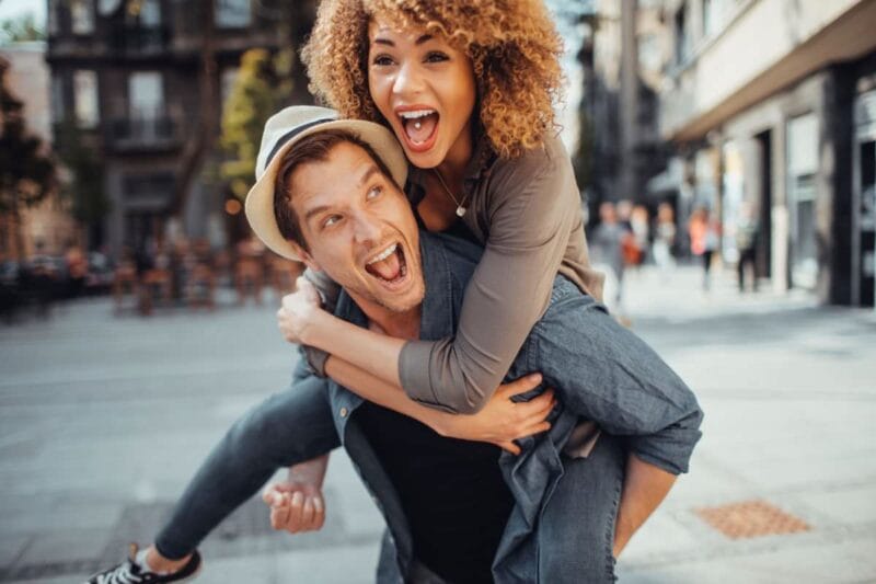 Ein glückliches Paar, das sich in der Stadt amüsiert, während der Mann die Frau auf dem Rücken trägt, lebendige Verlobungsfotos.