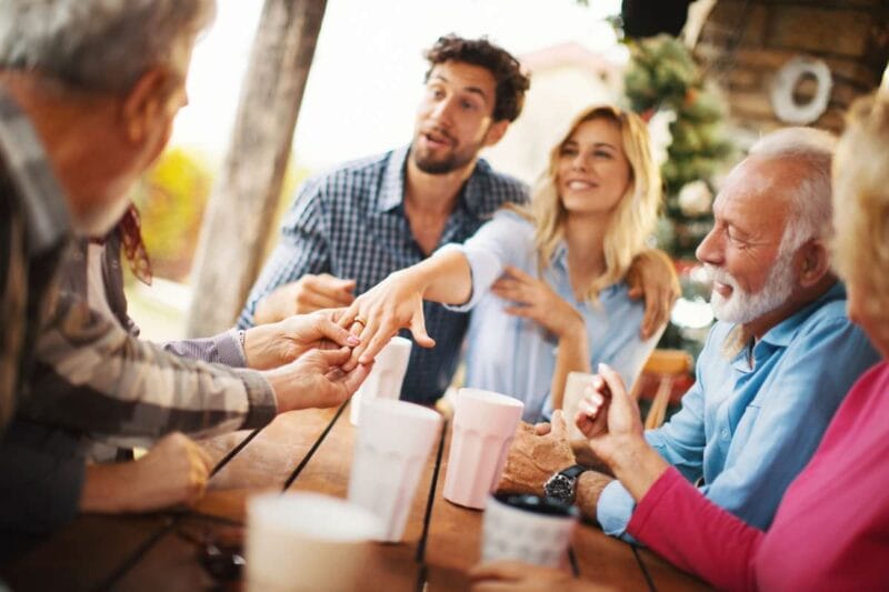 Glückliches Paar zeigt den Verlobungsring bei einer Verlobungsparty ihren Familienmitgliedern.