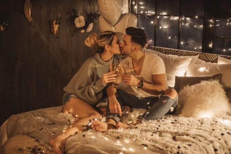 verliebtes Paar sitzt auf einem gemütlichen Bett und stößt mit Champagner an, umgeben von warmen Lichtern – eine sinnliche Darstellung der Sprache der Liebe